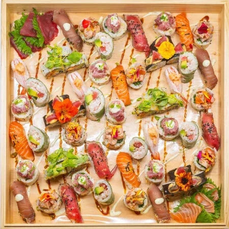 Un plateau en bois soigneusement disposé avec un assortiment de rouleaux de sushi, de nigiri, de sashimi et de garnitures, y compris des fleurs et des verdures comestibles.